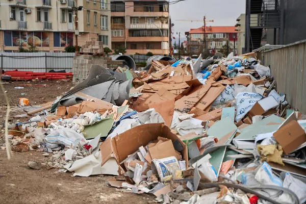 A pile of construction debris, including cardboard, plastic, and metal, is scattered at a site with buildings in the background. Efficient junk removal services are essential to clear this clutter swiftly and effectively.