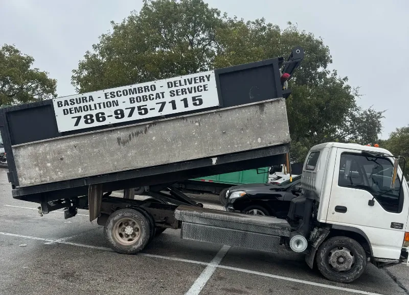 A white dump truck with raised bed is parked in a lot. It promotes trash pickup, waste management, demolition, and delivery services, featuring a phone number on the side. Trees stand tall in the background.