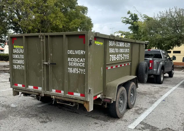 A green delivery and demolition trailer with a contact number is parked in a parking lot beside a truck, surrounded by trees.