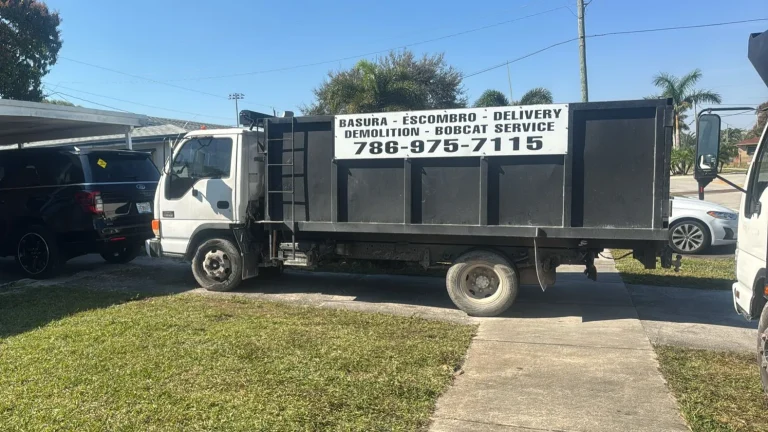 Black dump truck with signage for trash, debris removal, delivery, demolition, and bobcat service, parked on a driveway surrounded by vehicles and grass.