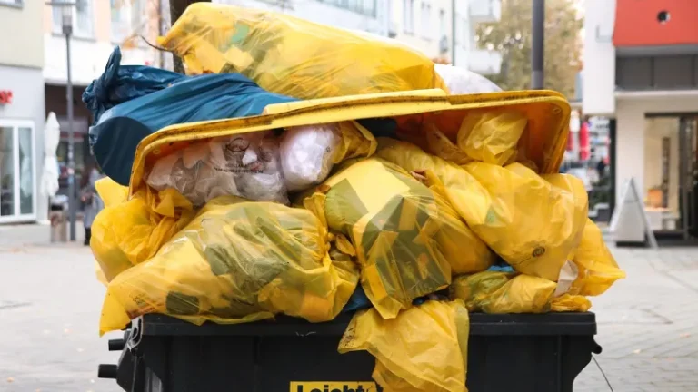 Overflowing dumpster with yellow trash bags, some spilling onto the ground, in an urban setting with buildings and trees in the background.