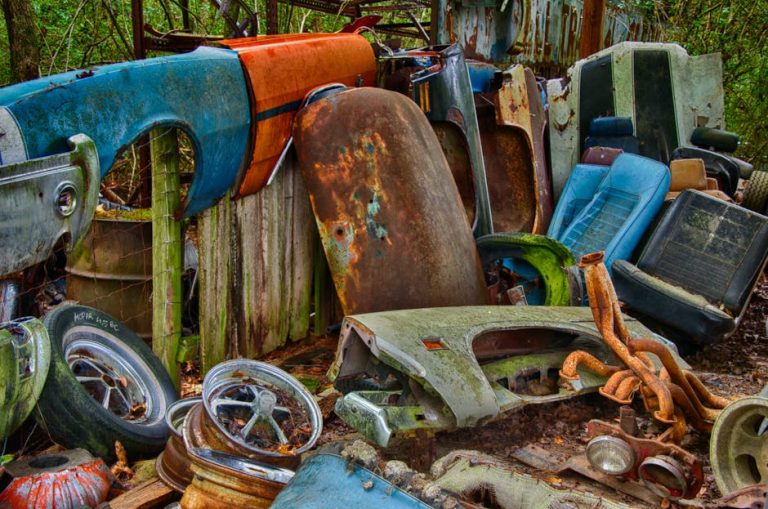 Amidst a cluttered outdoor setting surrounded by greenery lies a pile of discarded car parts—seats, doors, and tires. Consider booking junk removal with your local junk hauler to declutter efficiently and finally restore harmony to this chaotic scene.