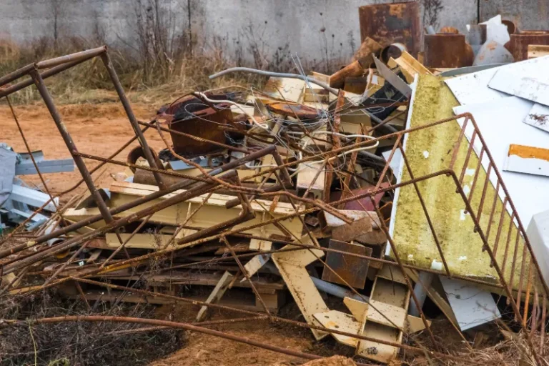 Pile of rusty metal scraps, including beams, rods, and sheets, on a dirt ground with dry grass and a concrete wall in the background.