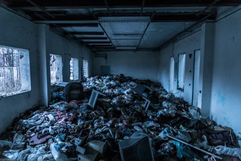 An abandoned room filled with piles of trash and debris, dimly lit by waning sunlight through dusty windows, shows signs of neglect and cries out for a professional trash removal service to restore its former glory.